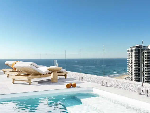 Poolside scene with wooden sun loungers, a small round table, and a stunning ocean backdrop, enclosed by clear glass railings under a bright blue sky.